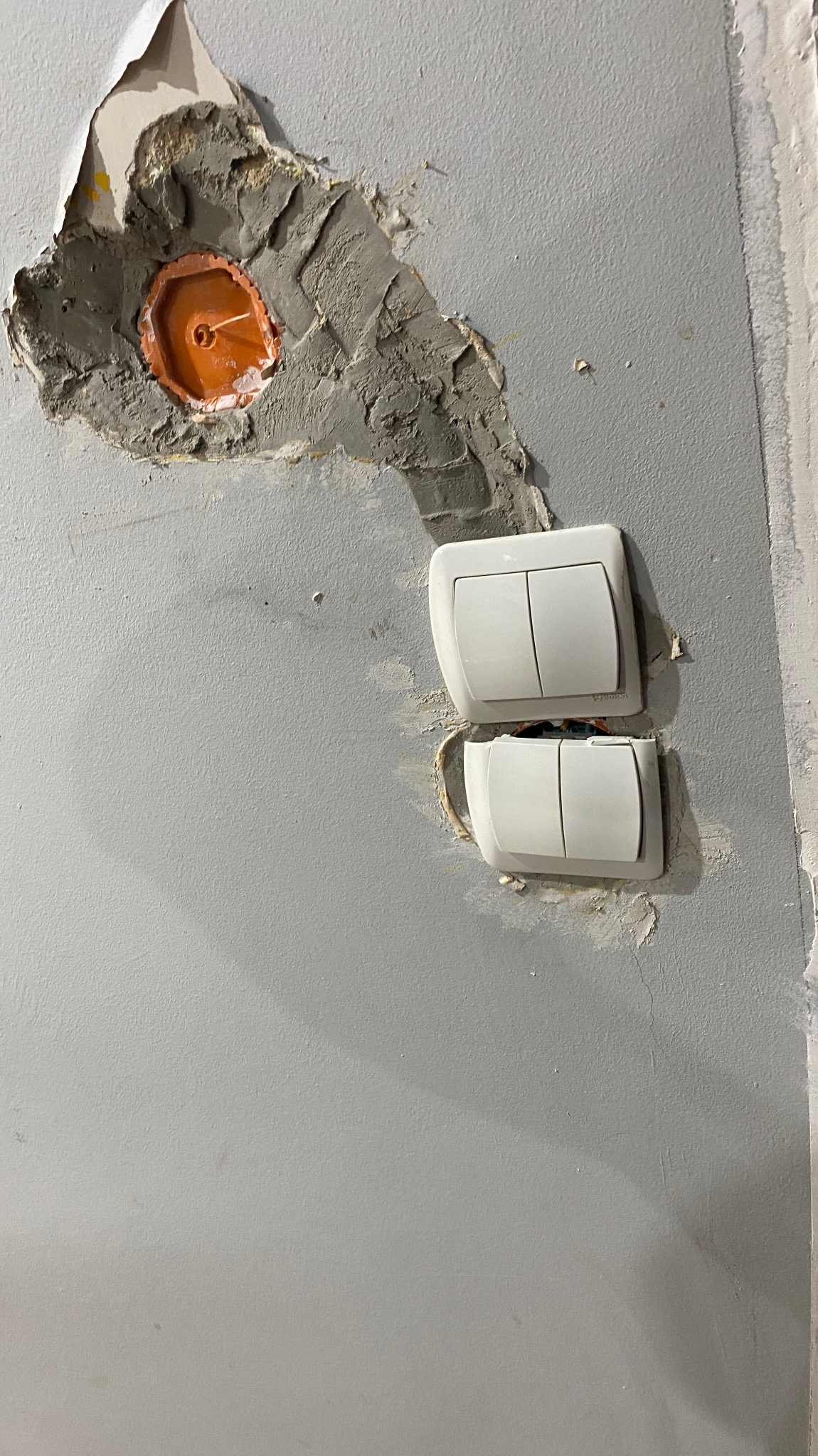 Dwa białe włączniki światła zamontowane na szarej ścianie, widoczne ślady po naprawach i niedoskonałości tynku wokół puszek elektrycznych.