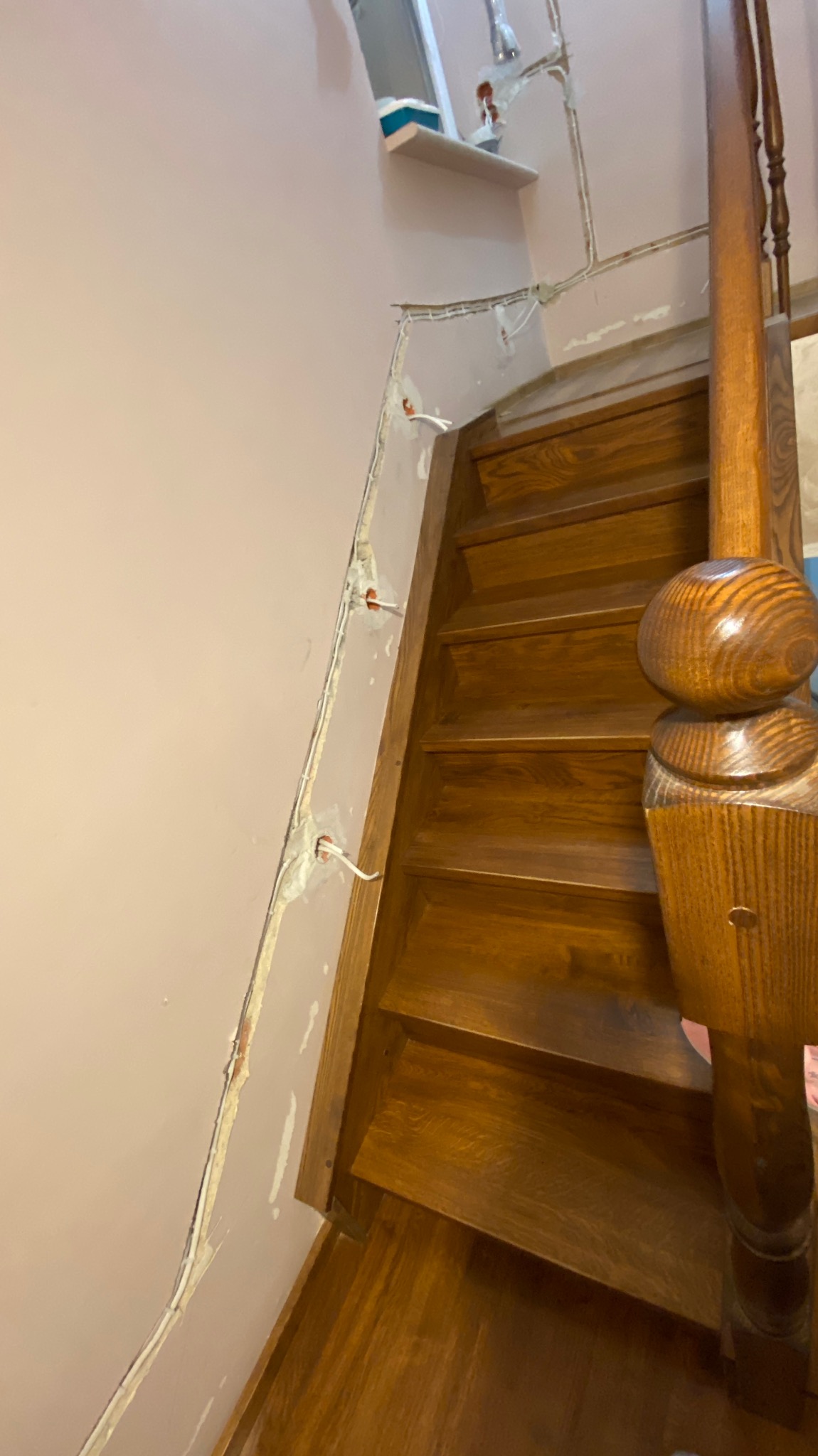 Wnętrze domu w trakcie remontu: drewniane schody z balustradą, różowa ściana z bruzdami na przewody elektryczne i wystającymi przewodami, widoczne ślady gipsu.