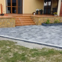 Szara kostka brukowa ułożona przed wejściem do domu z kamienną elewacją i schodami z płytek, obrzeża z jasnym paskiem, widok na trawnik.