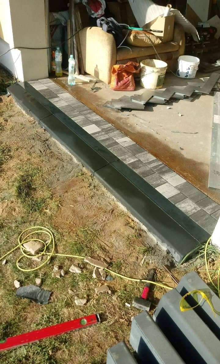 Układanie kostki brukowej: czarne obrzeże i szaro-biała kostka ułożona na trawniku, widoczne narzędzia i materiały budowlane.