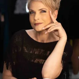 Portret kobiety z krótkimi blond włosami, ubranej w czarną bluzkę z frędzlami, z delikatnym uśmiechem i dłonią przy twarzy, eksponującej pierścionek na palcu.