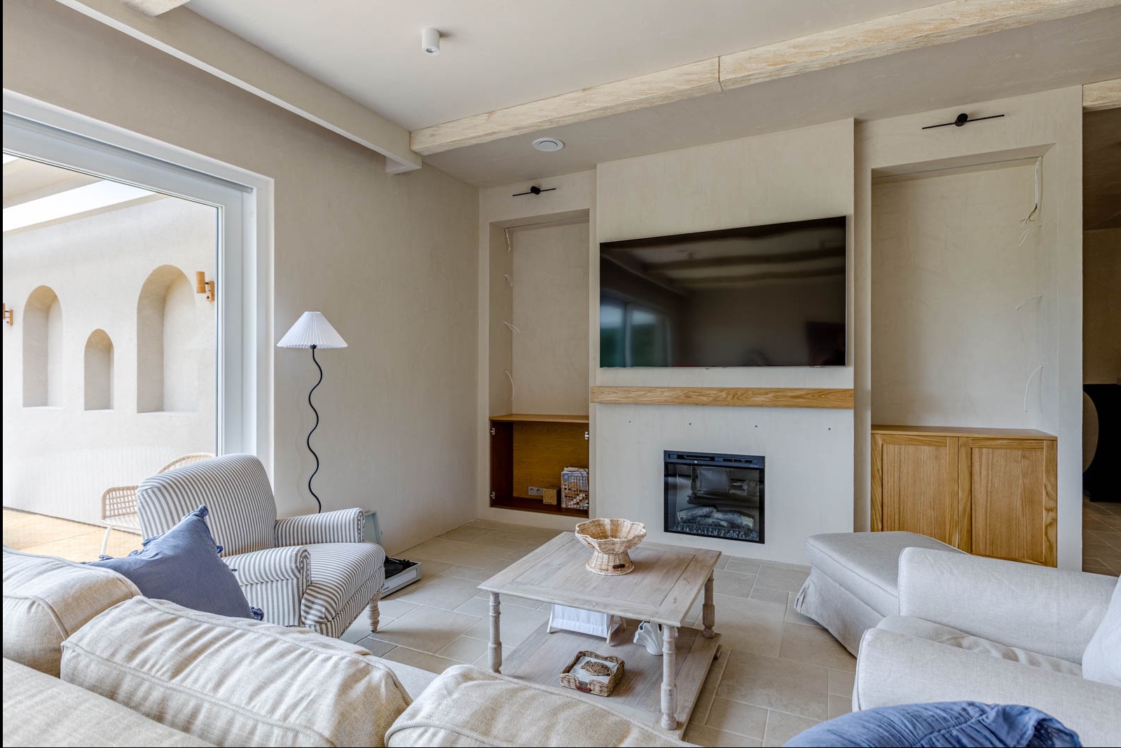 Jasne wnętrze salonu z beżowymi ścianami, kominkiem pod telewizorem i meblami w stylu skandynawskim. Widoczne okno z łukami i lampa ze plisowanym abażurem.