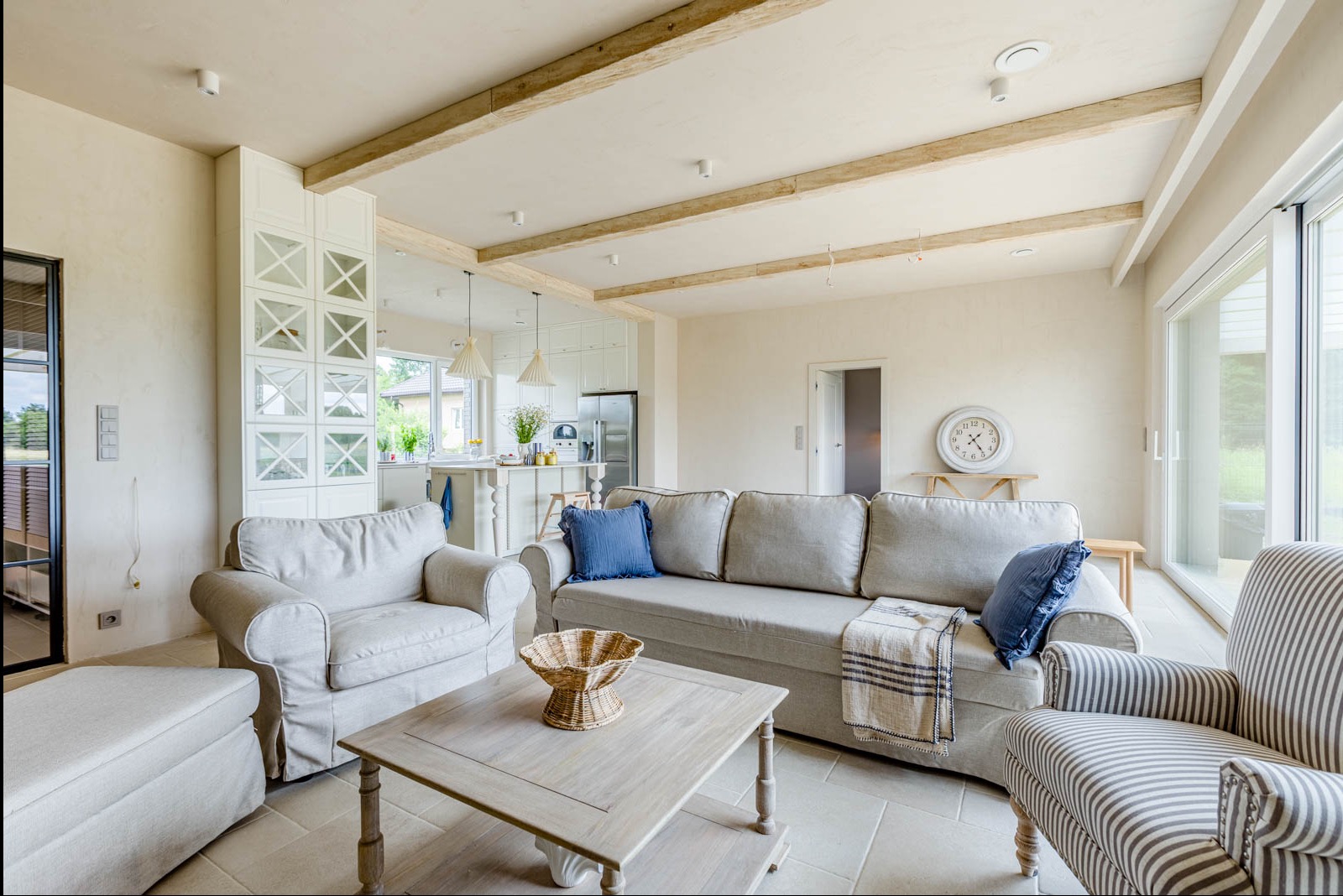 Jasne wnętrze domu z beżową sofą, drewnianymi belkami na suficie i otwartą kuchnią w tle. Widoczne krzesła, stolik kawowy i dekoracje.