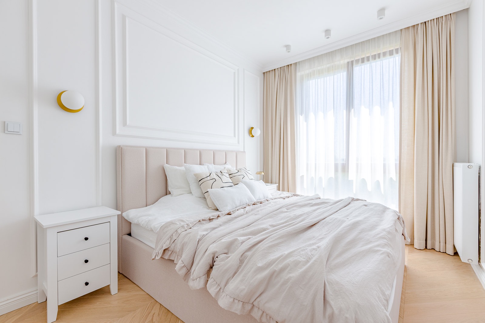 Jasna sypialnia z beżowym łóżkiem, białymi ścianami z dekoracyjnymi listwami i drewnianą podłogą. Okno z beżowymi zasłonami wpuszcza naturalne światło.