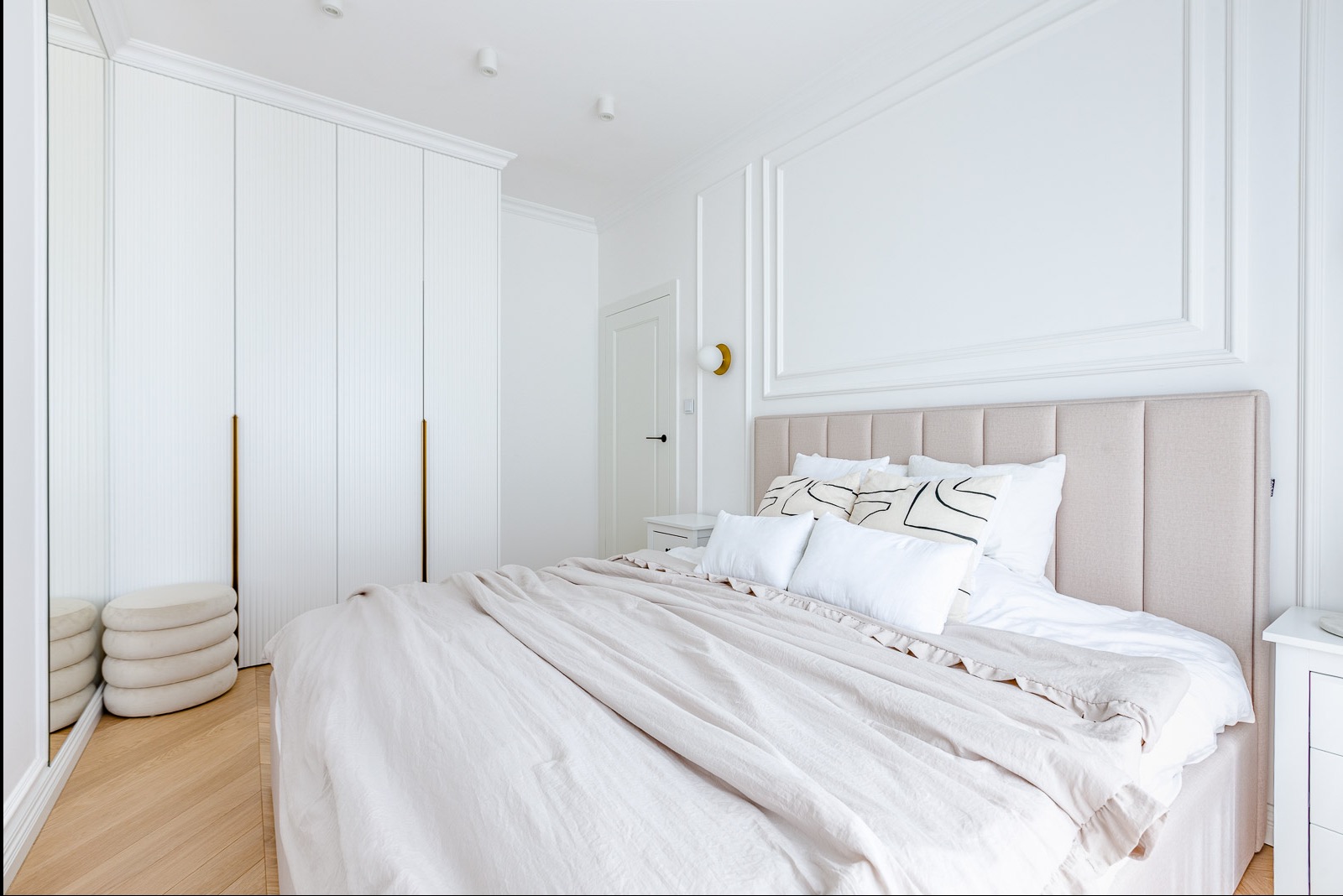 Jasna sypialnia w stylu minimalistycznym: beżowe łóżko z tapicerowanym zagłówkiem, biała pościel, szafa z pionowymi frezami i złotymi uchwytami, parkiet.