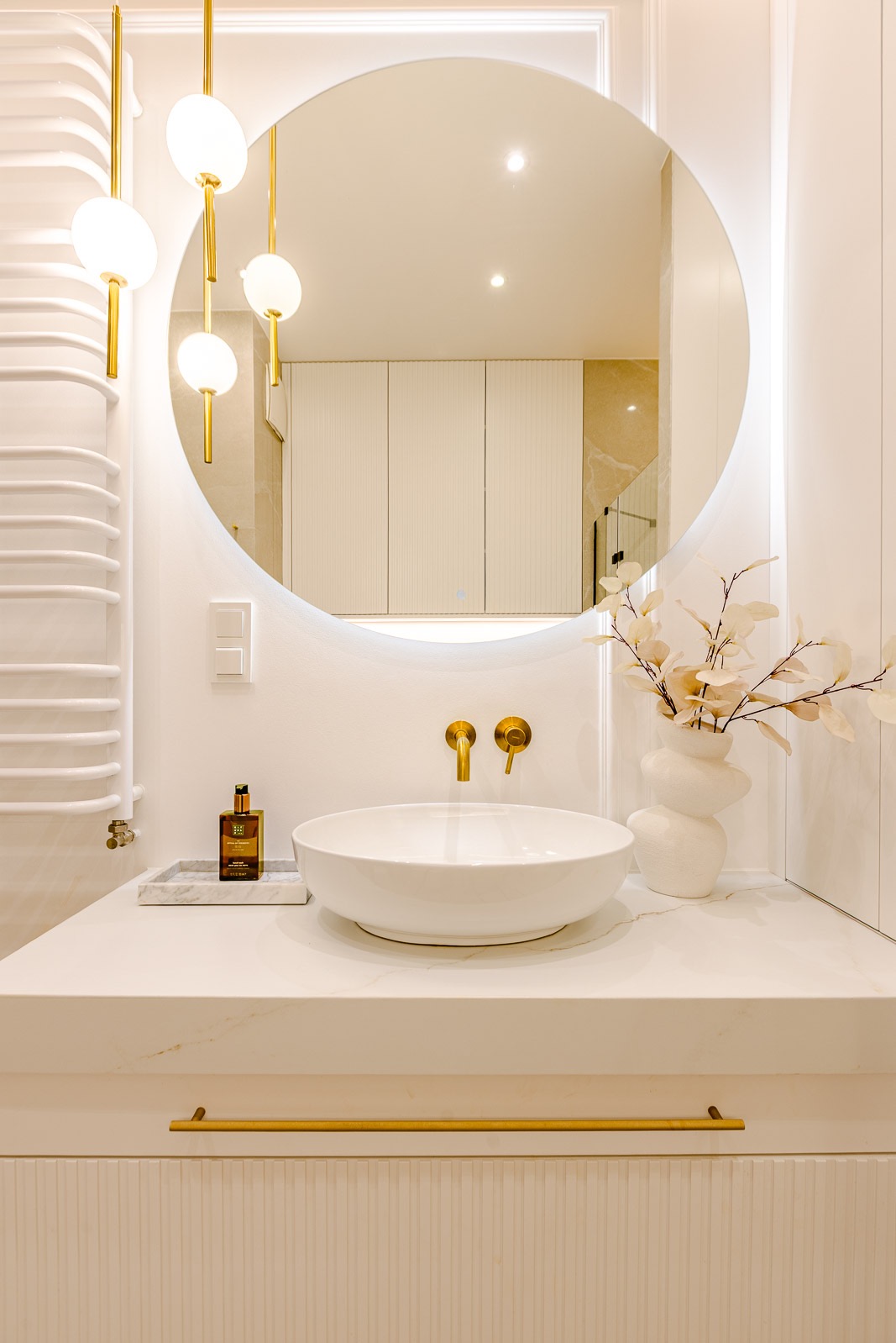 Elegancka łazienka w bieli i złocie: okrągłe lustro z podświetleniem, umywalka nablatowa, złota armatura, dekoracyjny wazon z gałązkami i designerska lampa.