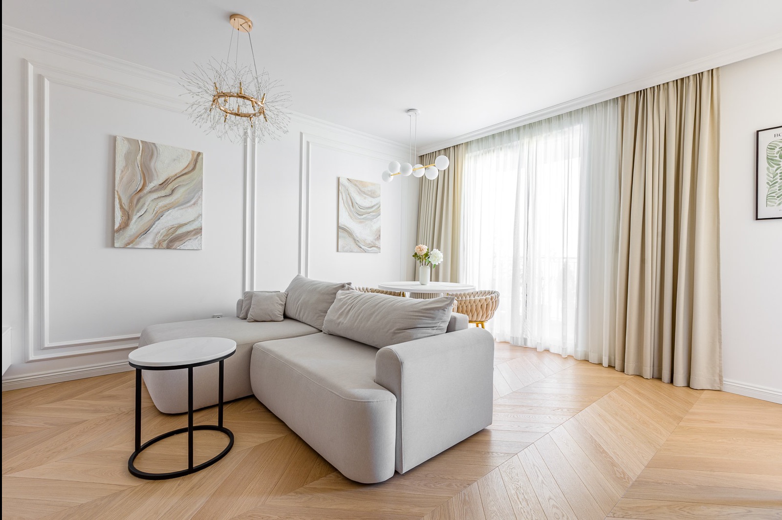 Jasne wnętrze salonu z narożnikiem, stolikiem kawowym i parkietem w jodełkę. Dekoracyjne obrazy na ścianach i designerskie lampy. Stonowana kolorystyka, naturalne światło.