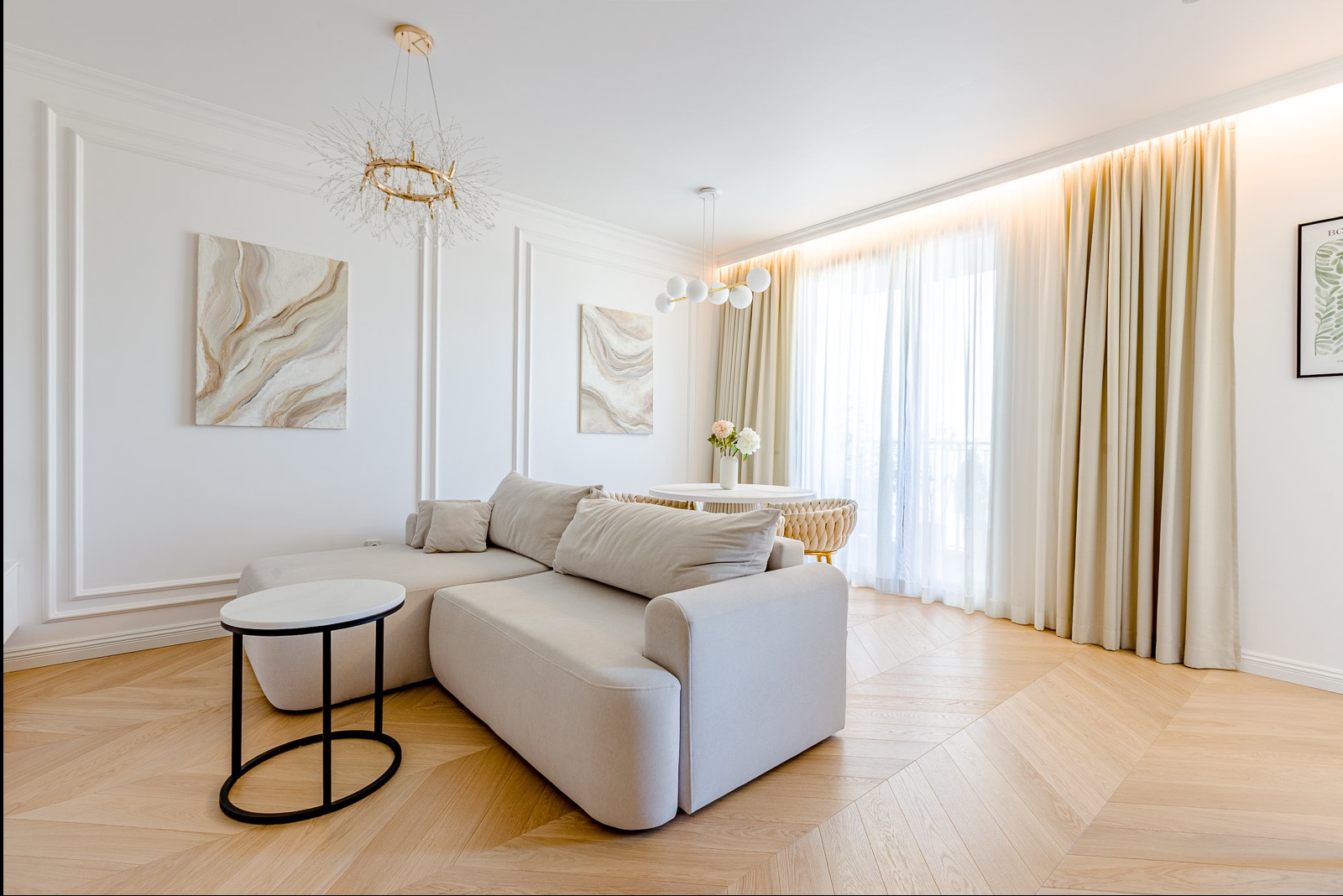 Jasny salon z beżową sofą narożną, okrągłym stolikiem i parkietem w jodełkę. Na ścianach obrazy w neutralnych barwach i dekoracyjne listwy. Całość doświetlona naturalnym światłem.