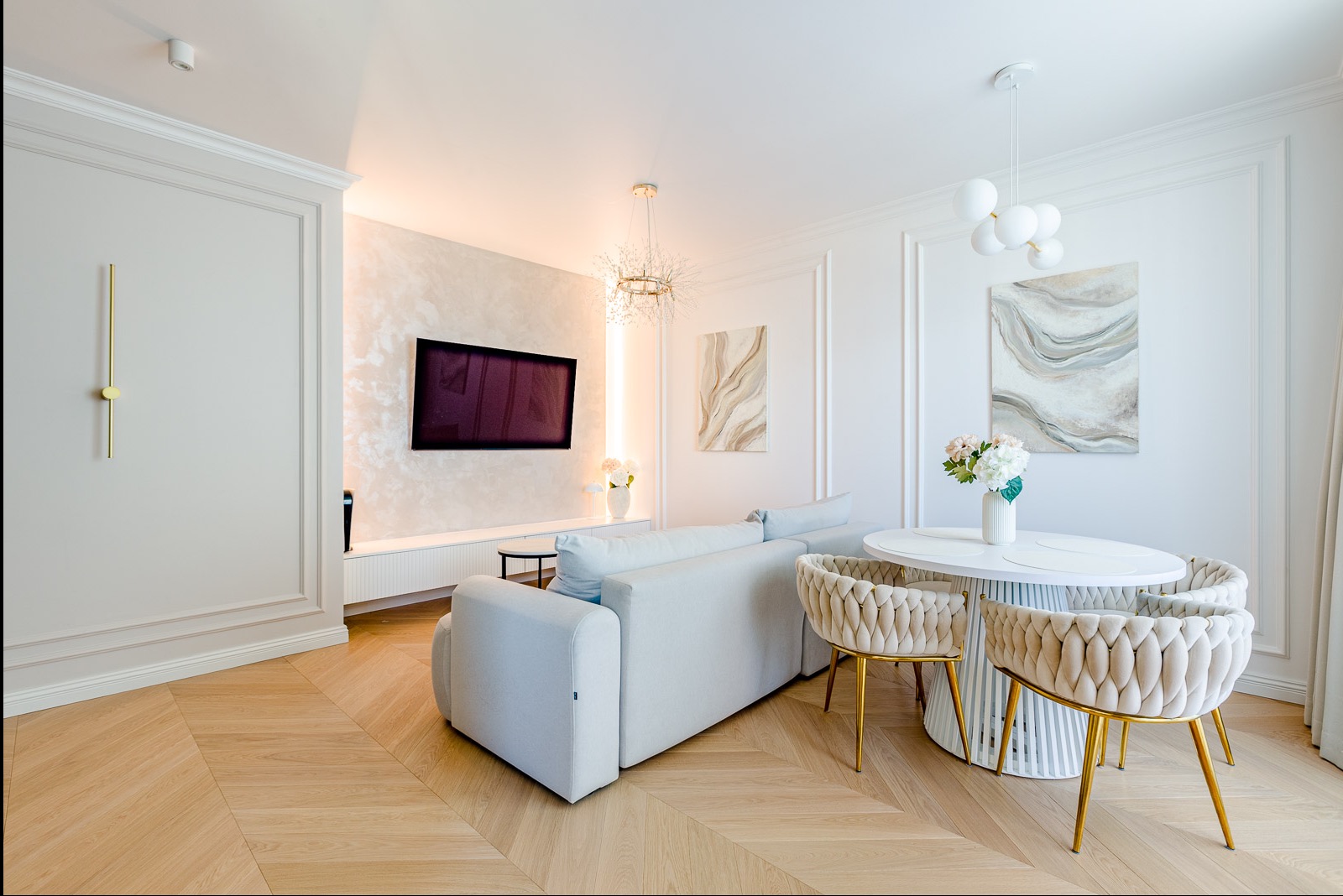 Jasne wnętrze salonu z parkietem w jodełkę, białą sofą i okrągłym stołem z krzesłami. Na ścianie telewizor i abstrakcyjne obrazy. Nowoczesne oświetlenie.