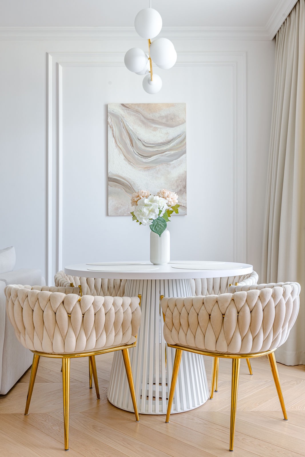 Jasne wnętrze jadalni: okrągły stół z kwiatami, beżowe krzesła z złotymi nogami, abstrakcyjny obraz na ścianie i designerska lampa sufitowa.