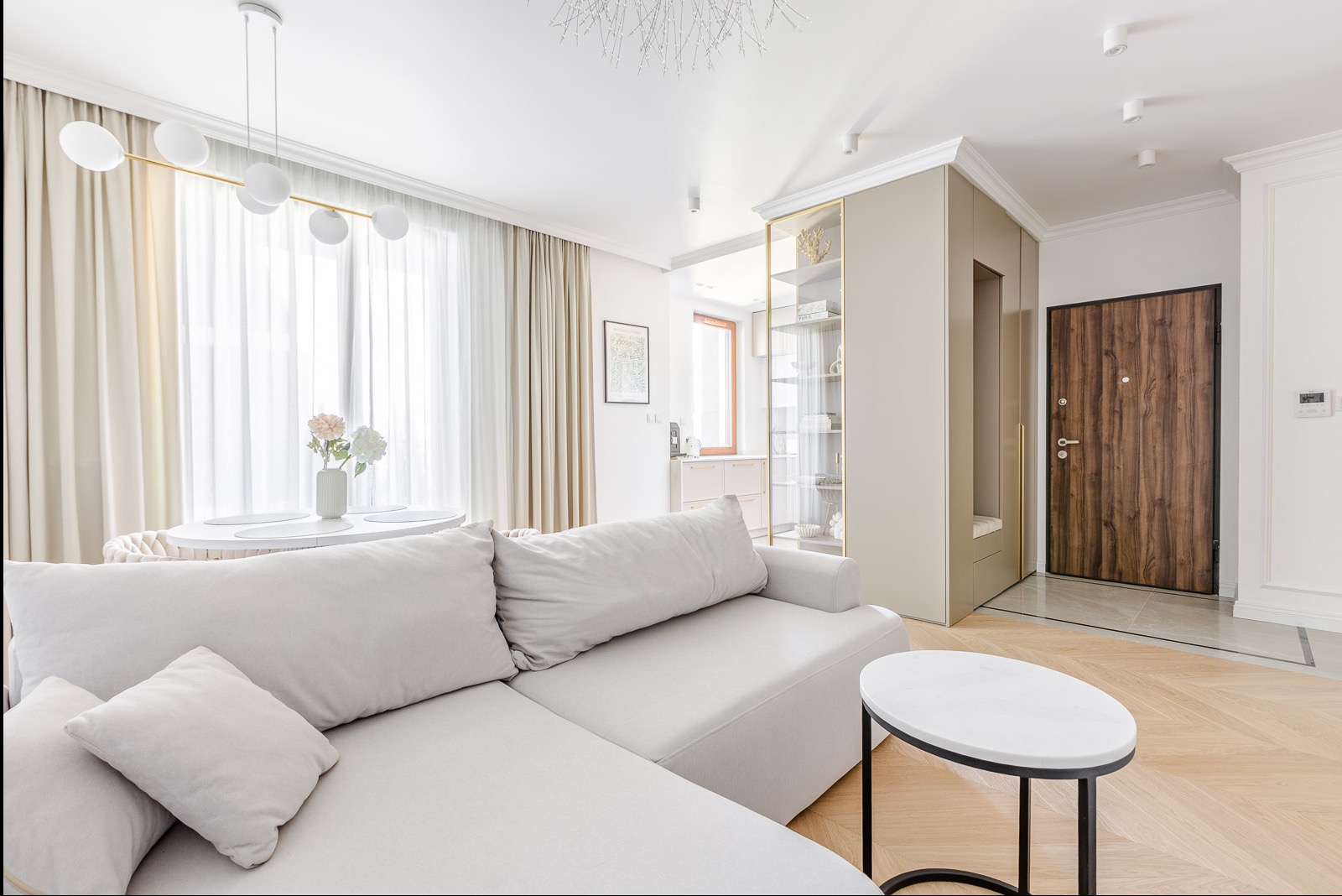 Jasne wnętrze salonu z narożną sofą, okrągłym stołem i drewnianymi drzwiami wejściowymi. Minimalistyczny design, parkiet w jodełkę, neutralna kolorystyka.