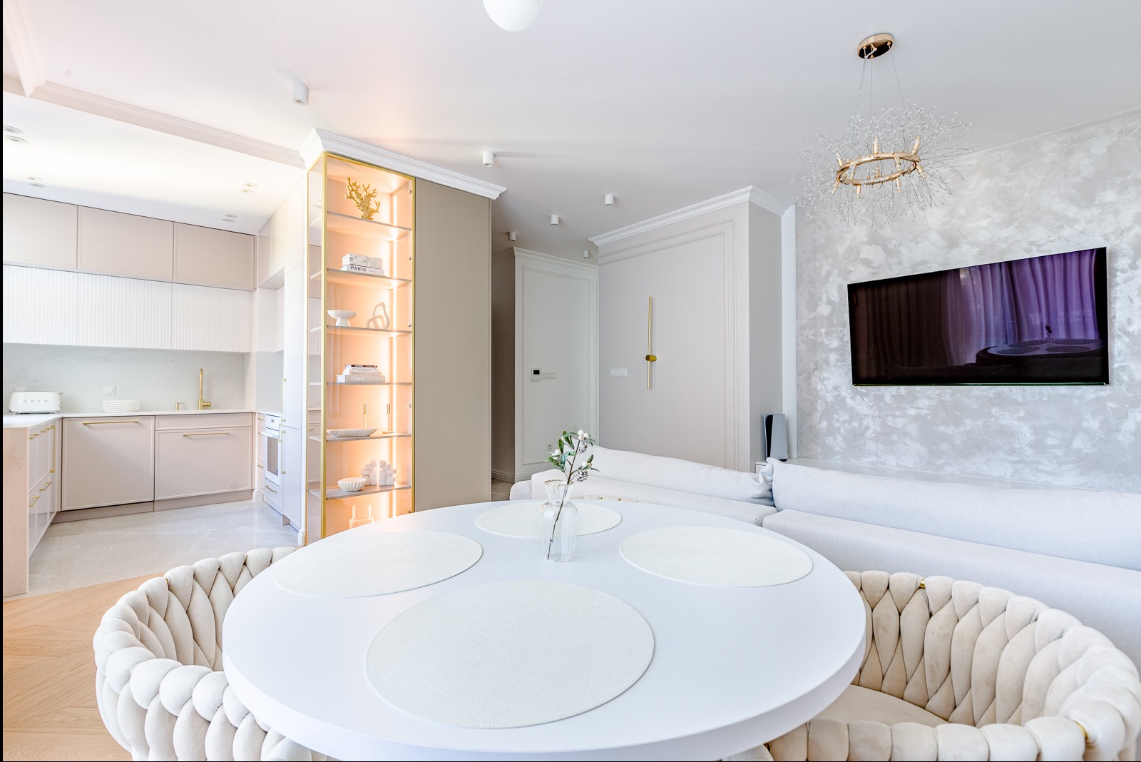 Jasne wnętrze: okrągły stół z krzesłami, sofa, regał z oświetleniem i telewizor. Minimalistyczny design, beżowo-biała kolorystyka, nowoczesne oświetlenie.