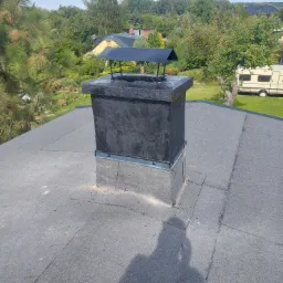 Szary dach pokryty papą z ciemnym, odnowionym kominem z czarnym daszkiem na tle zieleni drzew i zabudowań.