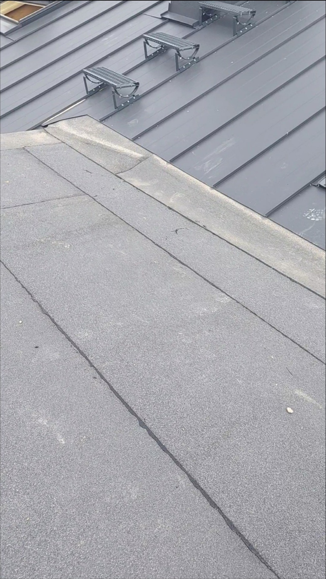 Fragment dachu z papy bitumicznej i blachy trapezowej z zamontowanymi stopniami kominiarskimi w kolorze grafitowym.