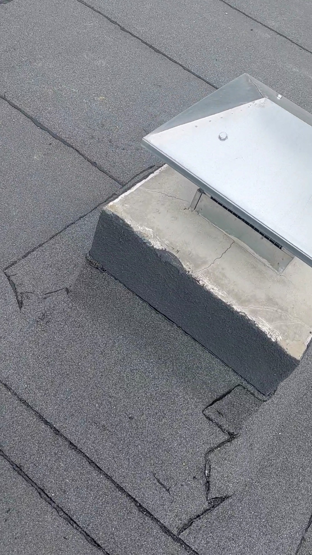 Szczegółowe ujęcie fragmentu dachu pokrytego papą z zamontowanym metalowym wywietrznikiem na betonowej podstawie. Widoczne pęknięcia w papie i na betonowej podstawie.