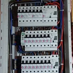 Rozdzielnica elektryczna z widocznymi bezpiecznikami, przewodami i listwami zaciskowymi, zamontowana w ścianie z ociepleniem z wełny mineralnej.