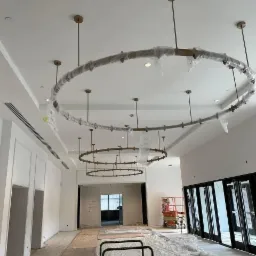 Montaż wiszących, okrągłych lamp w przestronnym, jasnym pomieszczeniu w trakcie prac wykończeniowych. Lampy zabezpieczone folią ochronną, widoczne narzędzia i podnośnik koszowy.