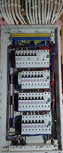 Rozdzielnica elektryczna z widocznymi bezpiecznikami, przewodami i listwami zaciskowymi, zamontowana w ścianie z ociepleniem z wełny mineralnej.