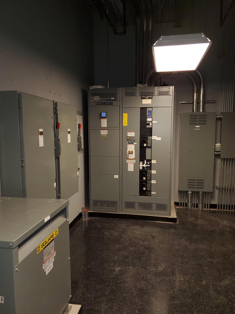 Szare rozdzielnice elektryczne Square D w pomieszczeniu technicznym z rurami na ścianach i posadzką z lastryko.