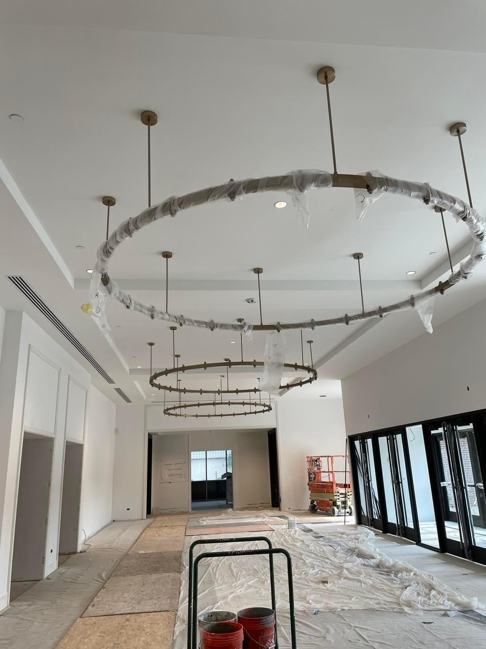 Montaż wiszących, okrągłych lamp w przestronnym, jasnym pomieszczeniu w trakcie prac wykończeniowych. Lampy zabezpieczone folią ochronną, widoczne narzędzia i podnośnik koszowy.