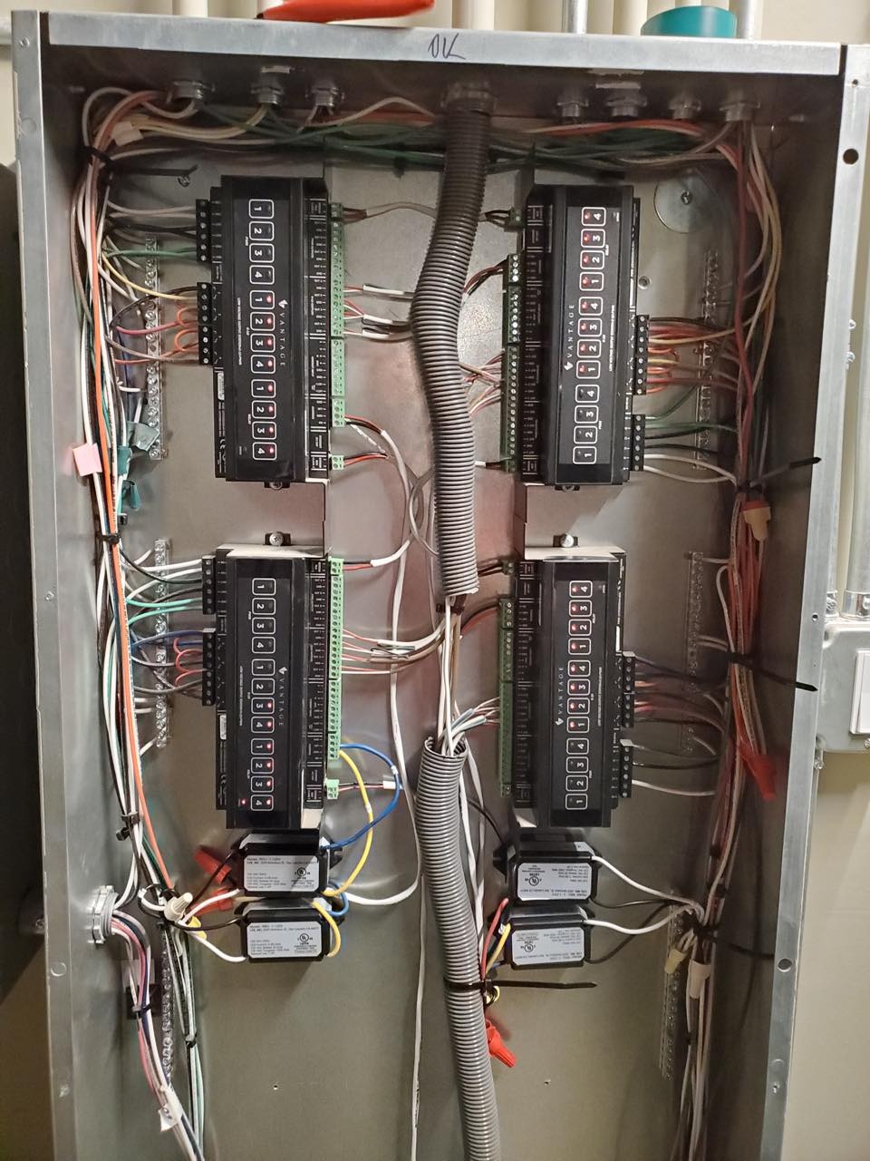 Otwarta metalowa skrzynka elektryczna z czterema czarnymi modułami sterującymi Vantage, licznymi przewodami o różnych kolorach podłączonymi do listew zaciskowych, oraz szarą karbowaną rurą ochronną...