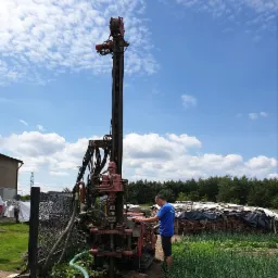 Wiertnica do studni głębinowej w trakcie pracy na działce, operator w niebieskiej koszulce przy panelu sterowania, widoczne przewody, rury i fragment ogrodu warzywnego.