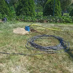 Niebieska rura wystająca z ziemi otoczona żwirem, żółty wąż i zwój czarnego kabla na trawie, w tle zielony żywopłot.