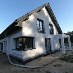 Nowoczesny dom jednorodzinny z białą elewacją, grafitową dachówką i czarnymi ramami okiennymi. Widoczna podbitka, barierki balkonowe i częściowo wykonane ocieplenie fundamentów. Na pierwszym planie...