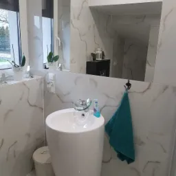 Biała, wolnostojąca umywalka z chromowaną baterią i szklanym elementem, obok pojemnik na śmieci, na ścianie płytki imitujące marmur, lustro i turkusowy ręcznik na haczyku.