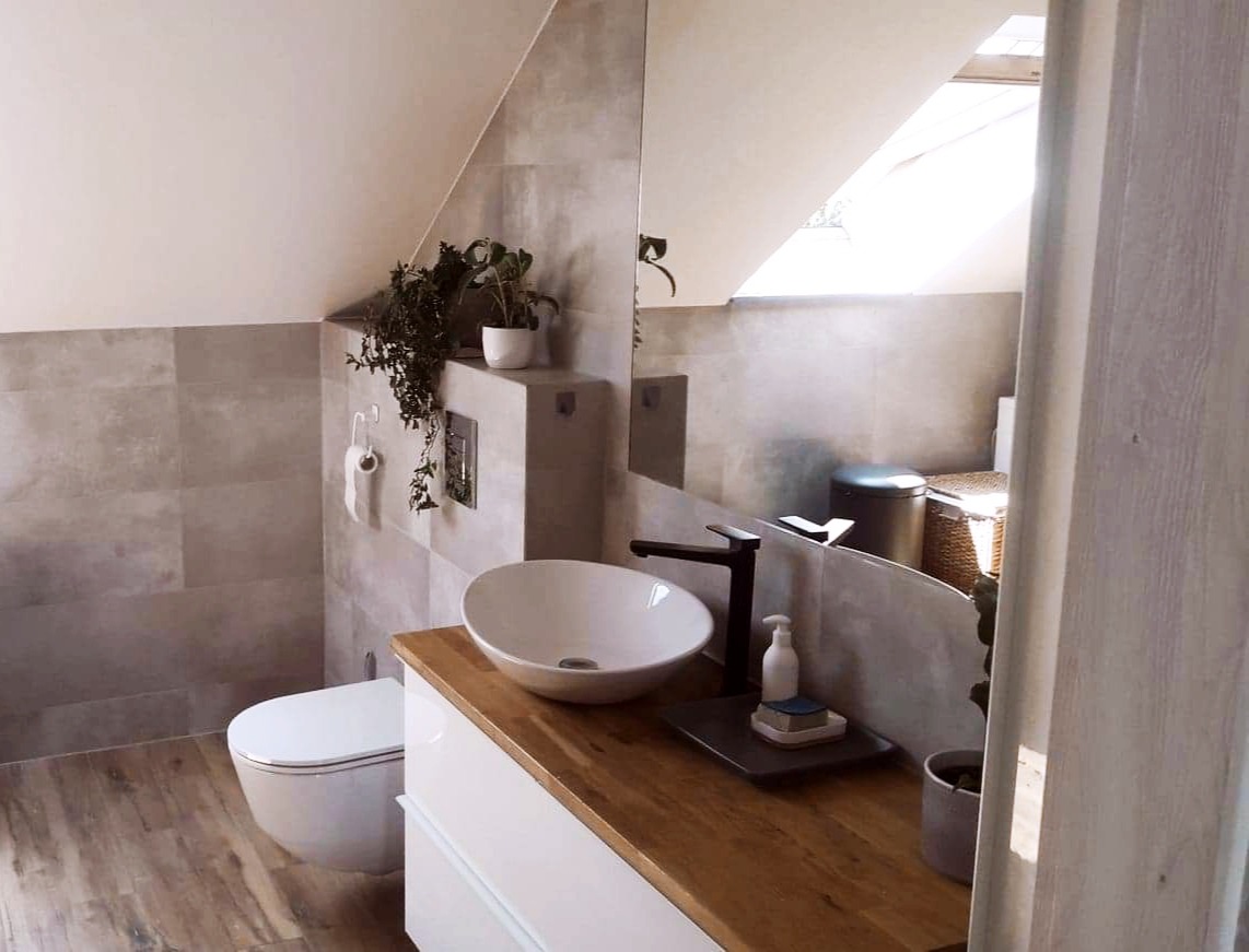 Nowoczesna łazienka na poddaszu z umywalką nablatową, drewnianym blatem, szarą płytką ścienną, wiszącą toaletą i roślinami doniczkowymi.