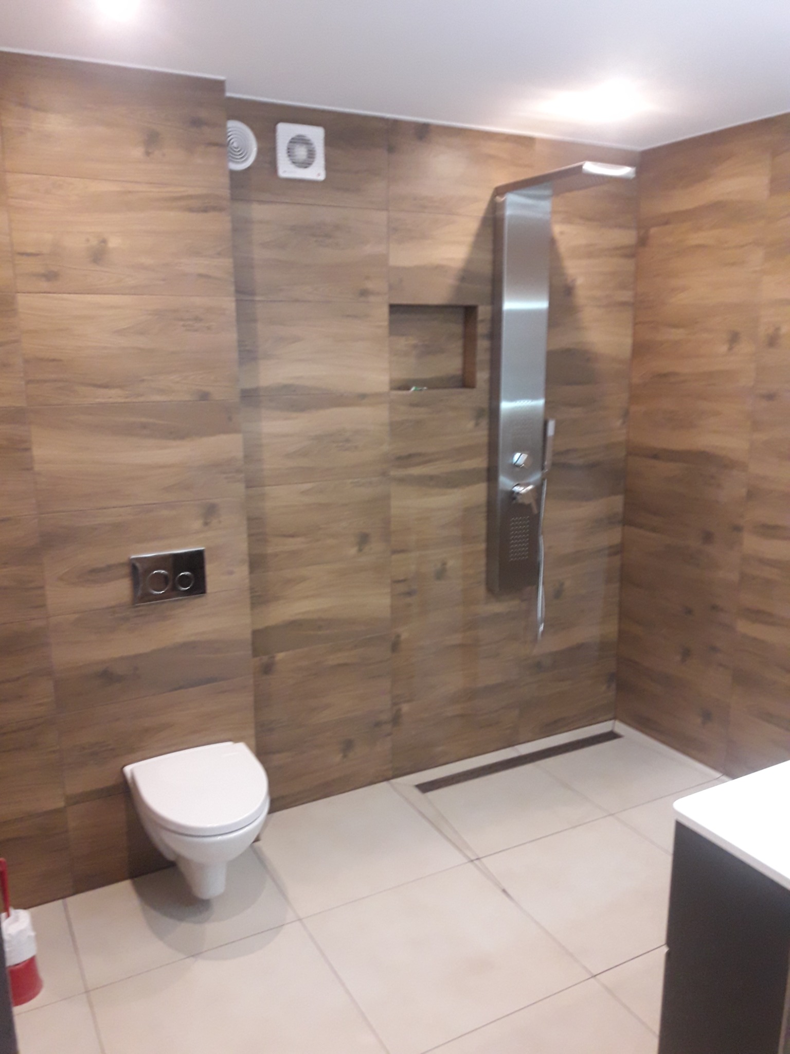 Nowoczesna łazienka z brązowymi płytkami imitującymi drewno, kabina prysznicowa z panelem natryskowym ze stali nierdzewnej, podtynkowa spłuczka WC z chromowanym przyciskiem i odpływ liniowy...