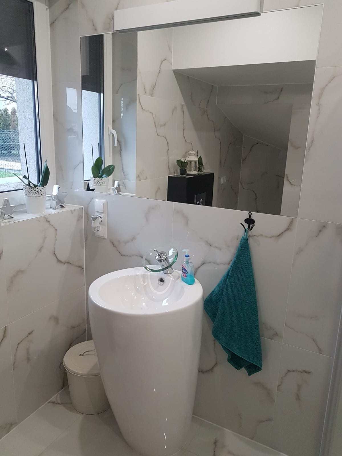 Biała, wolnostojąca umywalka z chromowaną baterią i szklanym elementem, obok pojemnik na śmieci, na ścianie płytki imitujące marmur, lustro i turkusowy ręcznik na haczyku.