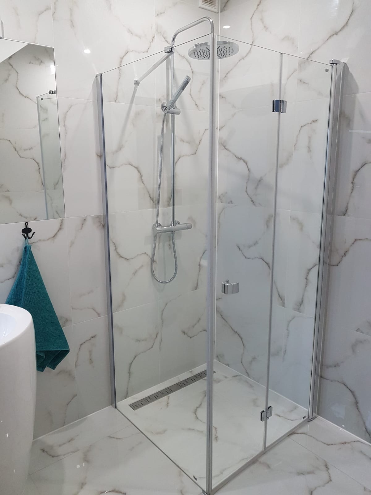 Nowoczesna kabina prysznicowa z deszczownicą i odpływem liniowym, wykończona białymi płytkami imitującymi marmur, z turkusowym ręcznikiem na haczyku.