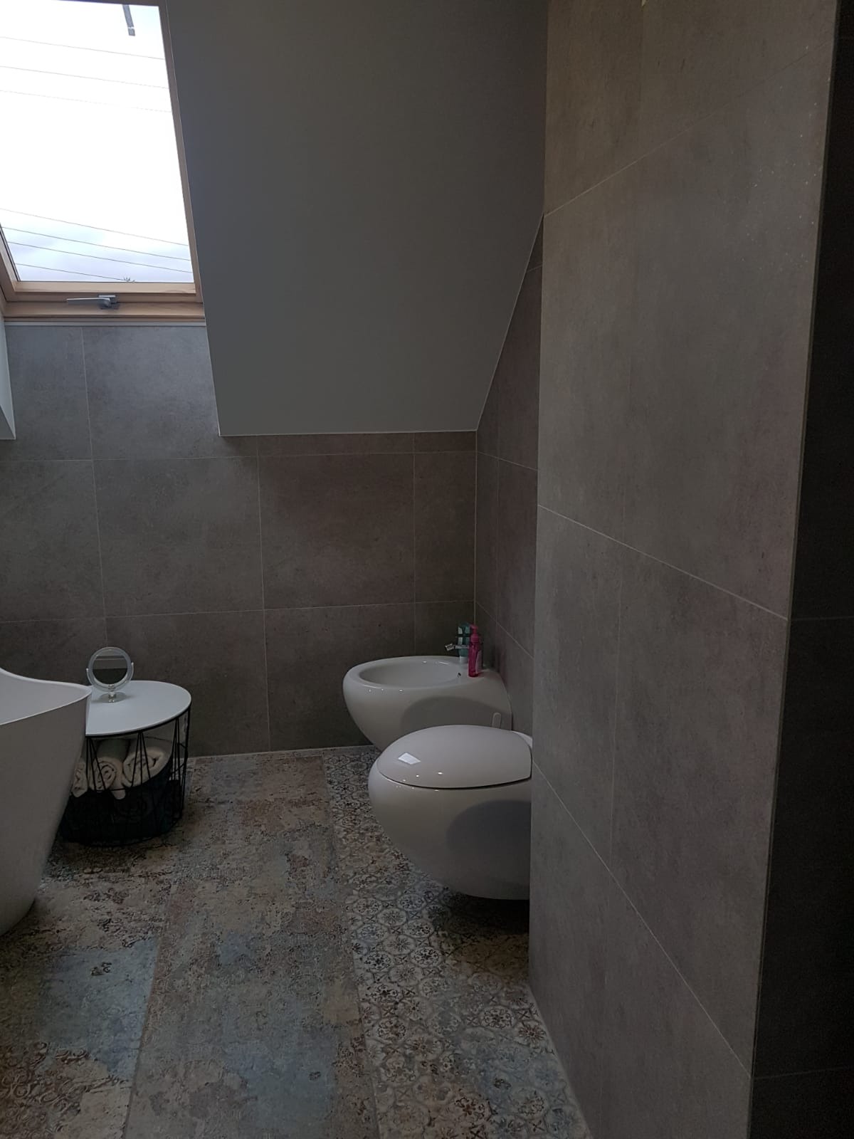 Szara łazienka na poddaszu z oknem dachowym, widoczne płytki na ścianach, toaleta, bidet, fragment wanny oraz stolik z lustrem.