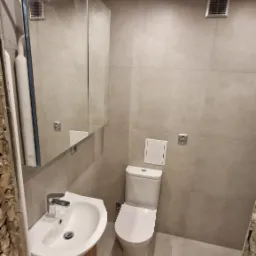 Nowoczesna, wykończona łazienka z umywalką na drewnianej szafce, toaletą i dużym lustrem na ścianie z szarych płytek.