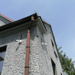 Dwie białe kamery monitoringu zamontowane na szarej, teksturowanej elewacji domu, widok z dołu, z brązową rynną i fragmentem dachu, na tle błękitnego nieba.