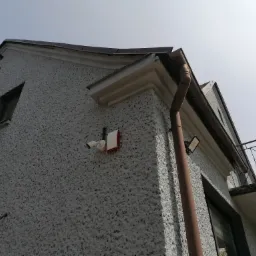 Kamera monitoringu i sygnalizator alarmowy zamontowane na szarej elewacji domu z widocznymi rynnami i balkonem, ujęcie z dołu.