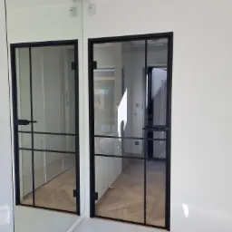 Szklane drzwi wewnętrzne z czarnymi szprosami, widoczne w lustrzanym odbiciu, z minimalistycznym designem w jasnym wnętrzu.