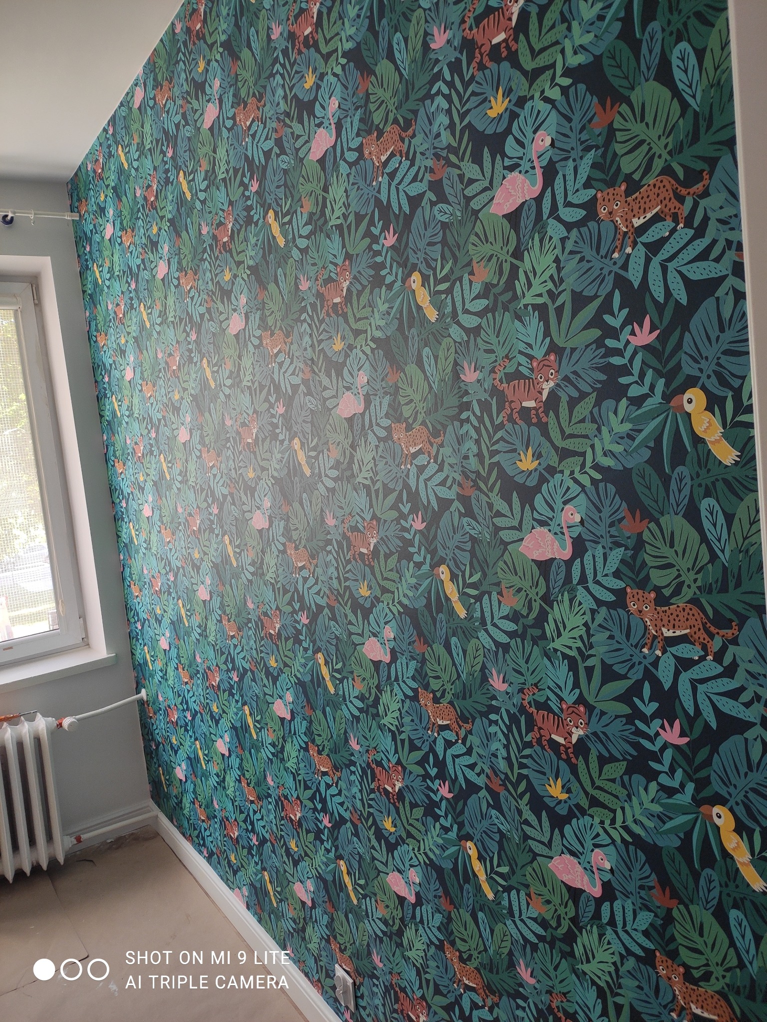 Ściana pokryta tapetą z motywem dżungli: liście, tygrysy, flamingi i papugi, widoczny fragment okna i grzejnika, podłoga zabezpieczona kartonem.