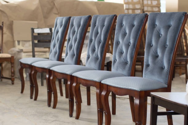 Rząd pięciu krzeseł z drewnianą ramą i tapicerowanym, szarym, pikowanym oparciem, ustawionych na betonowej posadzce, w tle widać meble przykryte folią.