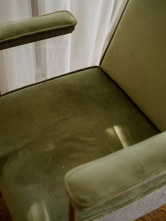 Fragment zielonego fotela tapicerowanego z widocznym siedziskiem, oparciem i podłokietnikiem, na tle białej zasłony.