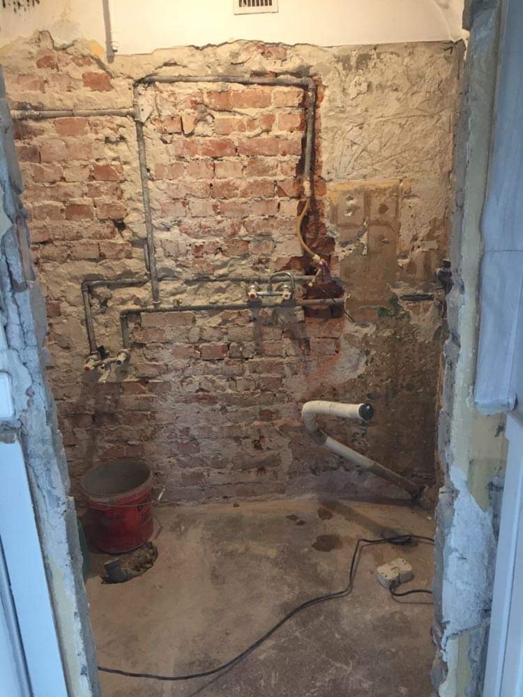 Odsłonięta ceglana ściana z instalacjami hydraulicznymi w trakcie prac przygotowawczych, widoczny fragment wentylacji pod sufitem, wiadro i kabel na podłodze.