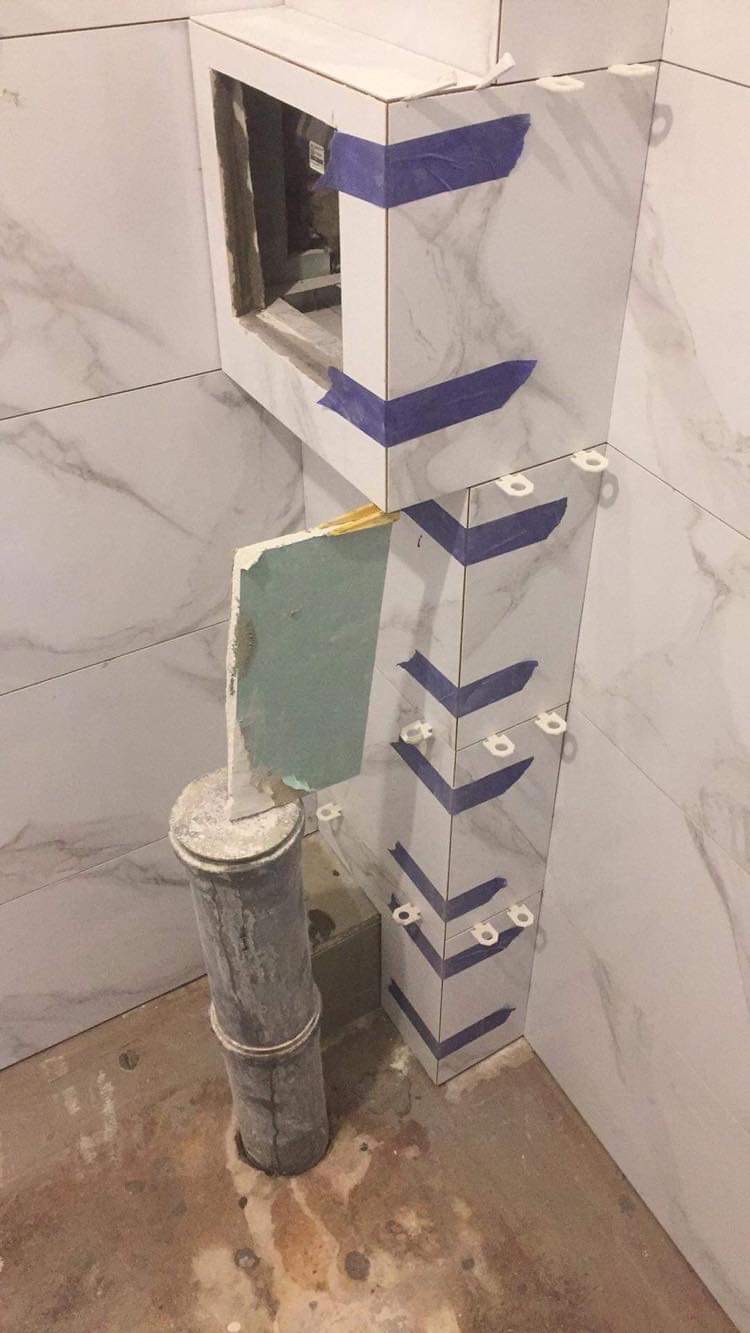 W trakcie prac w łazience: obudowa z płytek imitujących marmur, zabezpieczona niebieską taśmą, widoczne krzyżyki dystansowe, obok rura kanalizacyjna i kawałek płyty gipsowej.