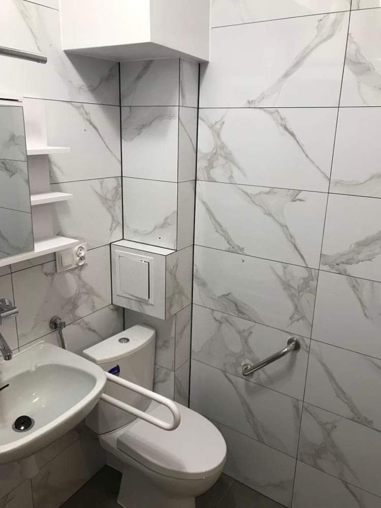 Wykończona łazienka z płytkami imitującymi marmur, biała ceramika sanitarna z uchwytami dla niepełnosprawnych, zabudowa nad toaletą z ukrytym zaworem.