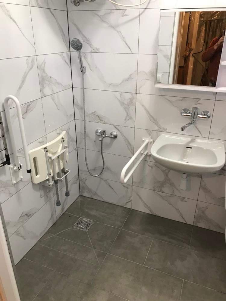 Wykończona łazienka w Busko-Zdroju z marmurowymi płytkami, wyposażona w składane siedzisko prysznicowe i uchwyty dla osób z niepełnosprawnościami, widoczny odpływ liniowy w podłodze.