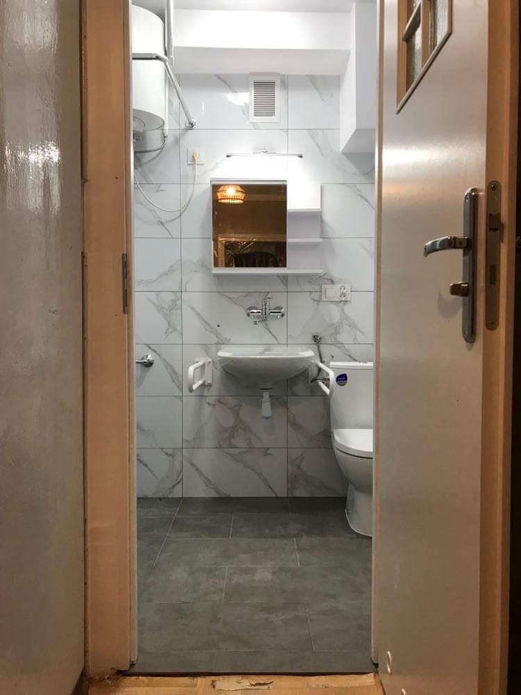 Widok na wykończoną łazienkę z marmurowymi płytkami, szarą podłogą i białymi sanitariatami, widzianą zza uchylonych drzwi.