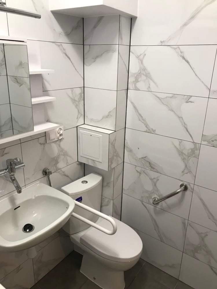 Nowoczesna łazienka z białymi płytkami imitującymi marmur, umywalka z chromowaną baterią, toaleta z uchwytem dla osób niepełnosprawnych oraz dodatkowy uchwyt na ścianie.