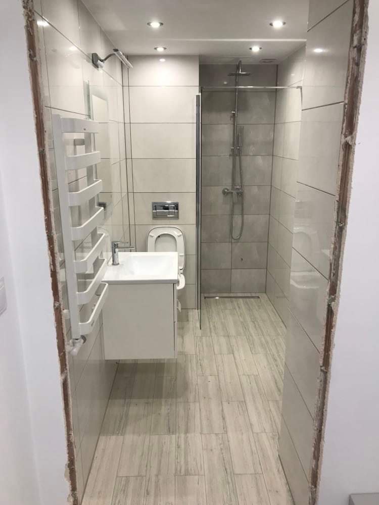 Nowoczesna łazienka z kabiną prysznicową typu walk-in, umywalką z szafką, grzejnikiem drabinkowym i płytkami imitującymi drewno na podłodze, widziana z perspektywy wejścia, z widocznymi surowymi...