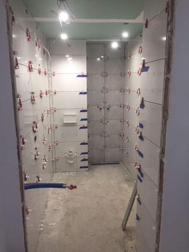 Wnętrze łazienki w trakcie prac wykończeniowych, widoczne białe płytki ceramiczne na ścianach, z użyciem systemu poziomowania płytek (klipsy i kliny), wystające rury instalacji wodnej, surowa podłoga.