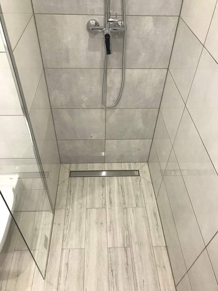 Kabina prysznicowa z odpływem liniowym, wykończona płytkami imitującymi drewno na podłodze i szarymi płytkami na ścianach.